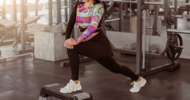 Spor giyinen bir kadın spor salonundaki bir platforma atlayıp spor rutinine odaklanıyor. Çevrede spor aletleri ve doğal ışık bulunmaktadır..