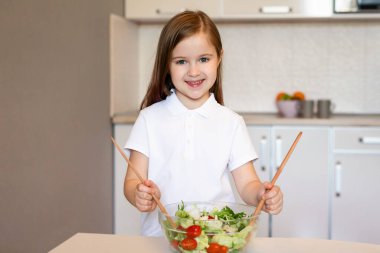 Küçük kız evde, modern mutfaktaki kameraya gülümseyen sebze salatası yapıyor. Mutlu Kadın Çocuk Karıştırma Yemeği Yemekleri Karıştırma Sebze İçindekileri Kasede Karıştırma. Sağlıklı Gıda ve Beslenme Konsepti