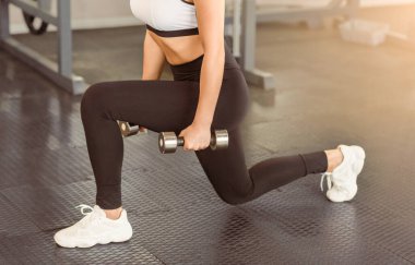 Bir kadın, iyi aydınlatılmış bir spor salonunda dambılları tutarken hamleler yaparak egzersiz yapar. Odaklanmış ifadesi, fitness ve güç eğitimine olan bağlılığını vurguluyor..