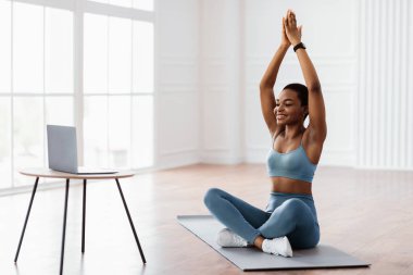 Genç bir Afrikalı Amerikalı kadın evde yoga yapıyor, minderde bacak bacak üstüne atmış oturuyor. Kollarını yukarı kaldırıyor, spor ve refahı teşvik ediyor, parlak, davetkar bir alanda..