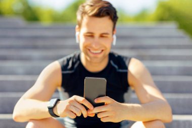 Alet edevatlı modern spor. Kablosuz kulaklıklı, akıllı saati ve akıllı telefonu olan gülümseyen adam parkta merdivenlerde oturuyor.