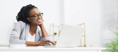 Afro-Amerikalı kadın dizüstü bilgisayara bakıyor ve gülümsüyor, ofiste, Panorama 'da yeni bir proje üzerinde çalışıyor, boşluğu kopyalıyor.