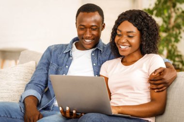 Güzel siyah çift evdeki oturma odasında kanepede dinlenirken dizüstü bilgisayar kullanıyor, birlikte fotoğraf izliyorlar.