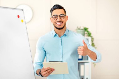 Genç bir Ortadoğulu işadamı beyaz bir tahtanın yanında dururken gülümsüyor ve onay veriyor. Elinde bir pano var. Şirketin ofisinde kendine güveni ve olumlu davranışları var..