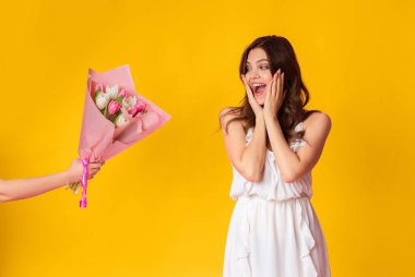 Memnun bir genç kadın, görünmeyen bir insandan bir buket çiçek alırken sevinç ifade eder. Parlak sarı arkaplan bu neşeli anda heyecanını vurguluyor..