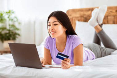 Asyalı Kadın, kredi kartı ve dizüstü bilgisayarını kullanarak internette alışveriş yapıyor ve evde yatıyordu. Online Satış ve İndirim. Seçici Odaklanma