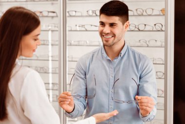 Bir adam optik mağazada bir kadına gözlük seçimi teklif ediyor. Çeşitli çerçeveler arasında konuşarak, konuşmalarla meşgul görünürler..