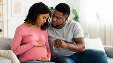 Bakım, hamilelik süresince çift desteği. Siyahi bir kadının hasta hissetmesini bekliyor. Kocası ona bir bardak su veriyor. İç mekan, boş yer. Afrikalı Amerikalı bir adam hamileliği boyunca karısına destek oluyor.