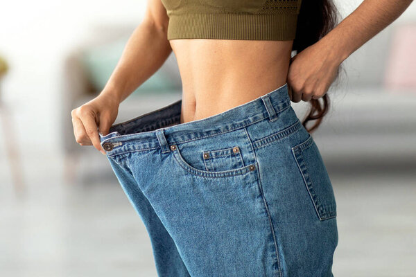 A young Indian woman shows off the fit of her oversized jeans after achieving her fitness goals. She embraces a healthy lifestyle through home workouts and balanced nutrition.