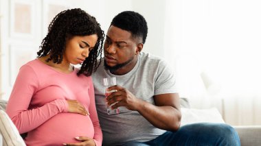 Bakım, hamilelik süresince destek. Siyahi kadın hasta hissediyor, koca bir bardak su veriyor, ev içi, boş alan.