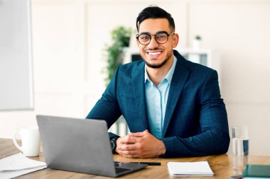 Genç bir Arap girişimci çağdaş bir ofiste masa başında otururken gülümsüyor. Resmi bir takım elbise giymiş, rahat bir tavırla dizüstü bilgisayarında profesyonel bir işe girmiş..