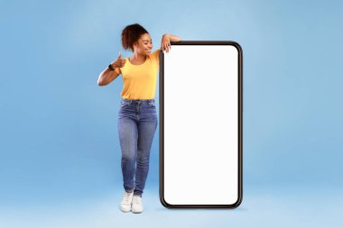 Kendinden emin bir Afro-Amerikan kadın boş bir akıllı telefonun yanında duruyor ve başparmağını kaldırıyor. Gülümsüyor, sarı gömlek ve kot pantolon giyiyor, bir anlık eğlencenin tadını çıkarıyor..