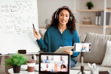 Esmer saçlı genç bir Orta Doğulu kadın online matematik dersi veriyor. Öğrenciler video çağrılarıyla uğraşırken, o kavramları açıklamak için beyaz tahta kullanıyor..