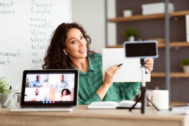 Esmer bir kadın rahat bir odadan çevrimiçi bir sınıfa liderlik ediyor, tabletinde öğretim materyalleri sergiliyor ve ilgili öğrencilere kavramları anlatıyor. Sahne etkili uzaktan öğrenmeyi yansıtıyor.