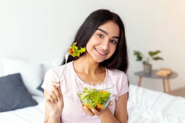 Genç bir Hintli kadın elinde taze salata tutarken gülümsüyor. Sıradan bir kıyafet giyiyor ve formda görünüyor. Evde sağlıklı bir yaşam tarzını benimsiyor..