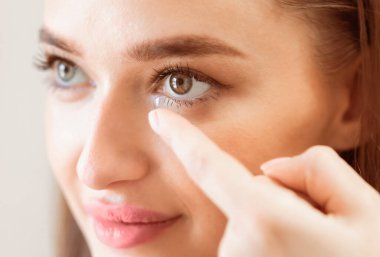 Genç bir kadın dikkatlice kontak lensini sağ gözüne yerleştirir, dikkatle odaklanır. Yumuşak ışıklandırma, titremeyen bir el ve sakin bir tavır takınırken onun özelliklerini ön plana çıkarıyor..