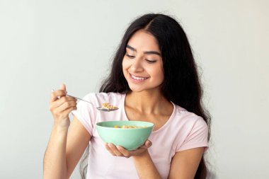 Genç bir Hintli kadın elinde bir kase mısır gevreği tutarken gülümsüyor, evde besleyici bir kahvaltı yapıyor. Sağlıklı bir yaşam tarzını ve kondisyon odaklı rutinini somutlaştırıyor, refahı arttırıyor..