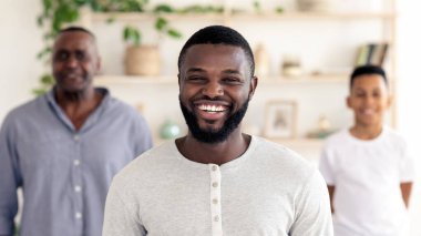 Açık gri gömlekli neşeli bir adam aydınlık bir odada geniş bir gülümseme sergiliyor. Aile üyeleri arkasında durup bitki ve dekorlarla dolu sıcak ve davetkar bir atmosfer yaratıyorlar..
