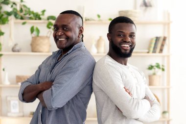 İki adam sırt sırta verip kolları bağlı gülümsüyor. Parlak oda modern dekor ve yeşillik içeriyor, neşeli bir atmosfer yaratıyor..