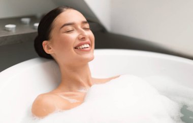 Gözlerini kapayan kadın köpükle banyo yapıyor suda yatıyor ve modern banyoda dinleniyor. Muhteşem Genç Bayan Banyo Yapan Spa Günü. Yurt İçi Rahatlama, Güzellik ve Sağlık