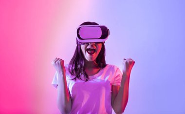 Bir bayan sanal gerçeklik teknolojisiyle neşe içinde etkileşime geçiyor, VR kulaklık takıyor. Parlak pembe ve mavi ışıklar çevresini aydınlatıyor, heyecanını ve nişanını yakalıyor..