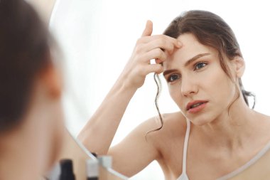 Üzgün sarışın kız alnına dokunuyor, aynaya bakıyor.