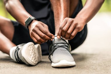 Spor giyim konsepti. Eğitimden önce ayakkabı bağcıklarını bağlayan, yolda oturan tanınmamış siyahi sporcunun yakın çekimi.