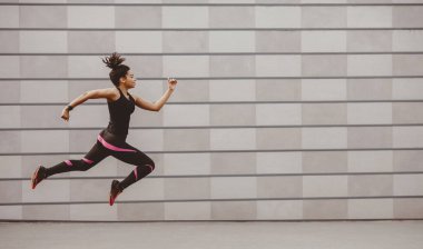 Mükemmel antrenman koşusu. Afrika kökenli Amerikalı bir kız, şık spor kıyafetleri içinde, yerin üstünde havada donarak, duvarın arka planında, yan görünüşünde, panoramasında, serbest alanda zıplıyordu.