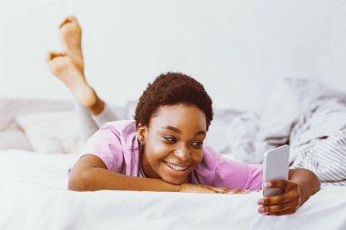 Yatak odasındaki yazışmalar. Gülümseyen Afrikalı Amerikalı kız akıllı telefondan bakıyor ve daktilo yazıyor.