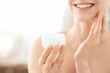 Yüz bakımı konsepti. Havluya sarılmış, krem sürerek tanınmaz hale gelmiş, gülümseyen bir kadın. Genç bayanların ellerinde yüz kremi olan kavanozlar, evde sabahları duştan sonra yüzüne nemlendirici sürüyorlar.