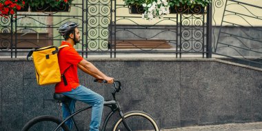 Kurye mal teslimatı için emirleri yerine getirir. Kasklı bir adam ve büyük sarı sırt çantalı bir adam yazın şehirde bisiklet sürüyor.