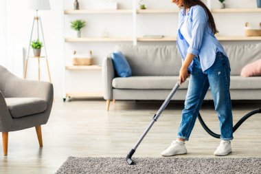 Genç bir kadın modern oturma odasındaki halıyı temizlemekten zevk alır. Günlük giysiler giyerken elektrikli süpürge kullanıyor. Pencereden güneş ışığı geçiyor. Boşluğu kopyalıyor.