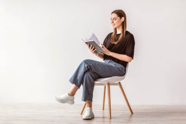 Uzun kahverengi saçlı beyaz bir kadın modern bir koltukta rahatça oturur ve bir kitap okumaya dalmıştır. Minimalist bir ortamda rahat bir yaşam tarzı sergiliyor..