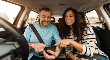 Kısa yol güzergahı. Lüks arabanın içinde oturmuş, akıllı telefonu işaret eden gülümseyen bir çiftin portresi, neşeli erkek sürücü hücresi, yeri gösteriyor, dijital harita uygulaması kullanıyor.