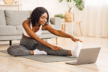 Zayıf siyah kız evde sabah egzersizi yapıyor, dizüstü bilgisayara bakıyor, boşluğu kopyalıyor.