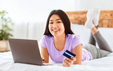 İnternetten alışveriş. Çinli bir kız kredi kartını dizüstü bilgisayarla alıyor ve internetten bir şeyler alıyor, kameraya gülümsüyor.