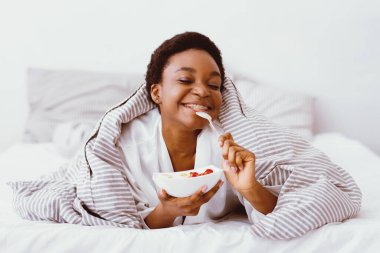 Sağlıklı kahvaltı. Memnun Afrikalı Amerikalı kız kaşığı dudaklarına dayayıp sağlıklı yiyeceklerle dolu tabağı tutuyor. Yatakta, boş alanda.