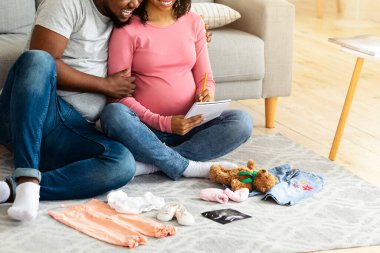 Neşeli hamile Afrikalı aile bebek kıyafetleri hazırlıyor ve liste yapıyor. Bebek doğurmaya hazırlanıyor. Oturma odasında yerde oturuyor.