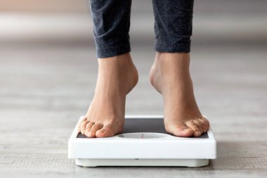 Genç bir Hintli kadın evde tartı terazisinde çıplak ayakla duruyor, sağlık ve spor hedeflerine odaklanıyor. Egzersiz yaparak dengeli ve sağlıklı bir yaşam tarzı sürdürmeyi hedefliyor..