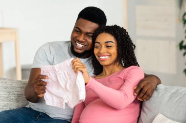 Gülümseyen siyah hamile çift pembe bebek kıyafetleri giyiyor, gelecekteki ebeveynliğin tadını çıkarıyorlar, ev içi, boş alan. Neşeli Afrikalı Amerikalı ebeveynler bebek şortlarına bakıp bebek bekliyorlar.