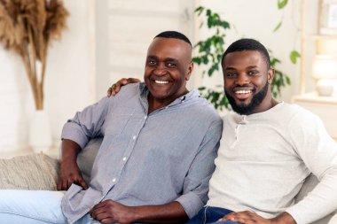 Bir baba ve oğul, rahat oturma odalarında keyifli bir anı paylaşırlar. Kanepede rahatça gülümserler. Etrafları bitkiler ve rahat dekorlarla çevrilidir..