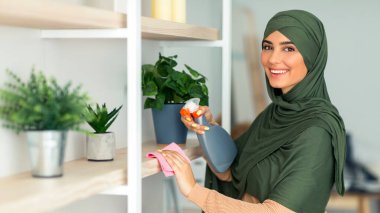 Genç Müslüman bir kadın, ev raflarını sprey şişesi ve bezle temizlemekle meşgul. Yeşil bitkilerle dolu yeri düzenlerken gülümsüyor, neşeli bir atmosfer sergiliyor, boşluğu kopyalıyor.