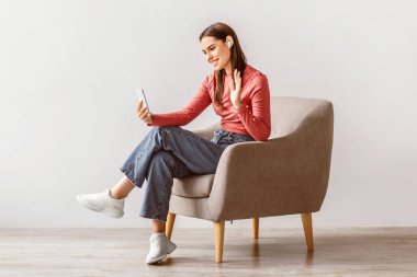 Esmer saçlı, beyaz bir kadın koltukta rahatça oturabilir. Akıllı telefonuyla yaptığı bir video görüşmesinin keyfini çıkarırken gülümseyip el sallıyor, rahat bir yaşam tarzı sergiliyor..