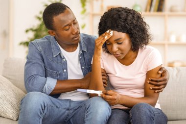 Kısırlık kavramı. Afrikalı üzgün bir adam üzgün karısına sarılıyor, hamilelik testi negatif, ev içi, boş alan var.