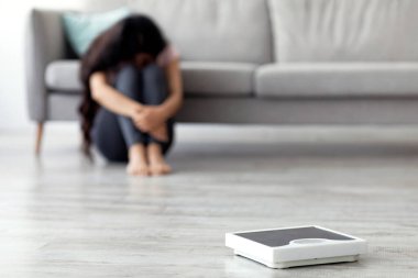 Genç bir Hintli kadın, spor ve sağlık hedeflerini düşünerek yerde oturuyor. Spor skalası yakınlarda duruyor. Huzurlu bir yaşam alanında sağlıklı bir yaşam tarzına olan bağlılığını vurguluyor..