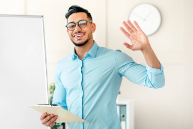 Genç bir Arap modern ofis ortamında duruyor. Elinde bir panoyla gülümsüyor ve el sallıyor. Meslektaşlarına fikirlerini sunmaya hazır..