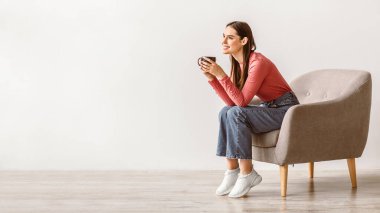 Esmer saçlı beyaz bir kadın şık bir sandalyede elinde fincanla oturuyor. Mutlu ve rahat görünüyor, minimalist bir ortamda huzurlu bir anı kucaklıyor..