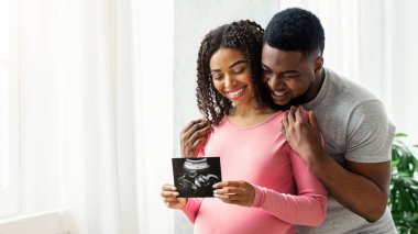 Gülümseyen hamile siyah çift, bebeklerinin ultrason görüntülerini gösteriyorlar. Neşeli afro-amerikan karı ve koca kucaklaşıyorlar ve ultrason embriyosu tutuyorlar, evde pencerenin önünde duruyorlar, boş uzay.