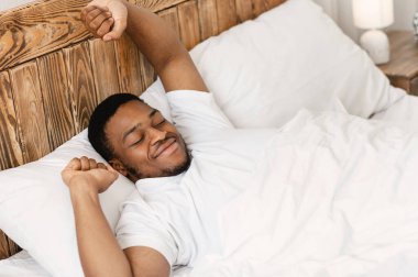 İyi sabahlar. Dinlenmis siyah adam ellerini uzatarak uyaniyor rahat bir yatakta uzaniyor, gözleri kapali bir sekilde gülüyor. İyi bir uykudan ve dinlenmeden sonra iyi uyuyan adam uyanıyor. Yüksek Açı Görünümü