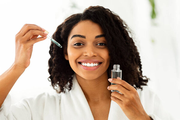 Skin Care Products. Smiling African Woman Holding Bottle With Moisturising Face Serum, Ready To Make Beauty Treatment After Shower